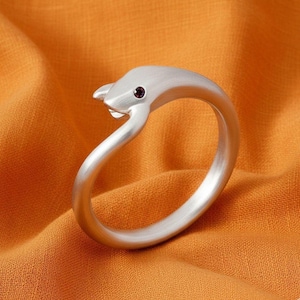 Könnte beinhalten: Ein silberner Ring in Form eines Delfins mit einem kleinen, dunkelroten Edelsteinauge. Der Ring liegt auf einem orangefarbenen Stoff. Der Körper des Delfins bildet das Ringband.