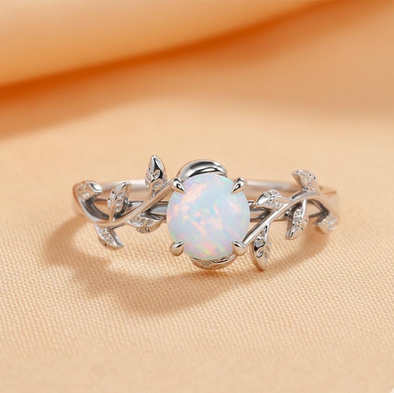 May include: A silver ring featuring an oval, opalescent gemstone. The band is adorned with a delicate leaf and vine design, with small accent stones. The ring is set against a soft, peach-colored background.