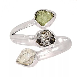 Puede incluir: Un anillo abierto de plata con tres piedras en bruto distintas engastadas en los extremos y el centro de la banda. Las piedras incluyen una gema verde claro translúcida, una piedra oscura con textura y un cristal transparente de corte irregular.