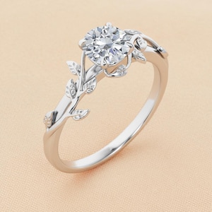 May include: A silver engagement ring with a large, round, clear gemstone. The ring features a delicate leaf design on the band and around the gemstone setting. The ring is set against a soft, neutral background.