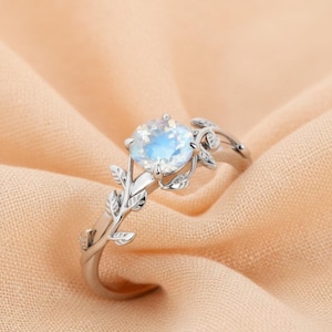 May include: A silver ring with a large, round, iridescent gemstone. The ring has a delicate vine design with leaves that wrap around the band and cradle the central stone. The ring is set against a peach-coloured fabric.