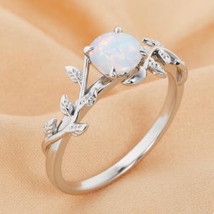 May include: A silver ring featuring a round, iridescent opal gemstone. The ring's band is adorned with a delicate leaf and vine design. The opal is set in a four-prong setting, and the ring has a polished finish.
