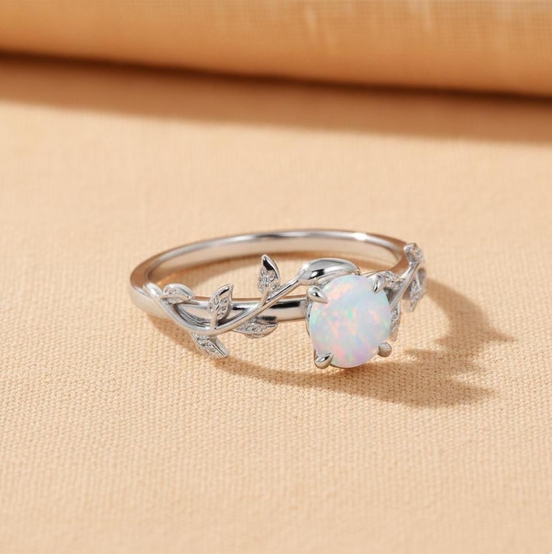 May include: A silver ring featuring a large, round, white opal gemstone. The ring band is adorned with a delicate leaf and vine design. The opal has a subtle, iridescent sheen. The ring is set against a neutral background.