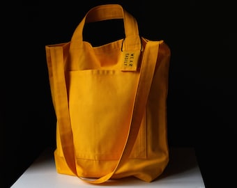 Può includere: Una borsa tote in tela gialla brillante con una grande tasca sul davanti. La borsa ha una tracolla lunga e un piccolo cartellino che dice "WEAR EASILY".