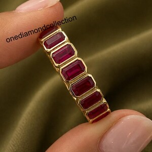 May include: A gold band ring featuring a series of rectangular, deep red gemstones set in a row. The text onediamondcollection is visible near the top of the ring. The design consists of multiple stones held in a continuous metal setting.