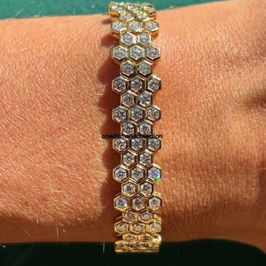 Könnte beinhalten: Ein goldenes Armband mit Wabenmuster, besetzt mit zahlreichen klaren, funkelnden Edelsteinen. Die sechseckigen Zellen sind mit glitzernden Steinen gefüllt. Das Armband wird am Handgelenk getragen.