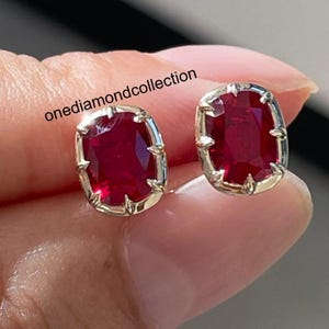 May include: A pair of stud earrings featuring cushion-cut, deep red rubies set in polished silver-toned metal prongs. The text onediamondcollection is visible across the image. Each ruby has a faceted surface that reflects light.