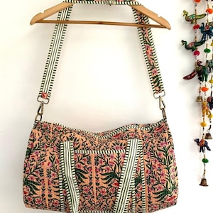 Puede incluir: Una bolsa de lona acolchada de color melocotón con estampado floral y correas a rayas verdes y blancas. La bolsa tiene una cremallera superior y dos asas.