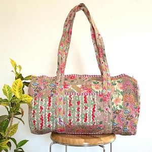 May include: A floral patterned quilted tote bag in pink, green, and beige. The bag features two handles and a rectangular shape. The design includes vertical stripes and a pink trim. The bag is placed on a wooden stool.
