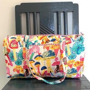Puede incluir: Una bolsa de lona blanca con un estampado de setas de colores. La bolsa tiene un patrón rosa, amarillo, azul y verde con una variedad de formas de setas. La bolsa tiene dos correas y un cierre de cremallera.