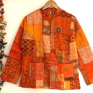 Könnte beinhalten: Eine orangefarbene Patchwork-Jacke mit Stehkragen und langen Ärmeln. Die Jacke besteht aus verschiedenen Stoffen mit Mustern in Orange, Rot und Gelb. Sie hat zwei Vordertaschen und wird mit Knöpfen geschlossen. Sie hängt an einem Holzbügel.