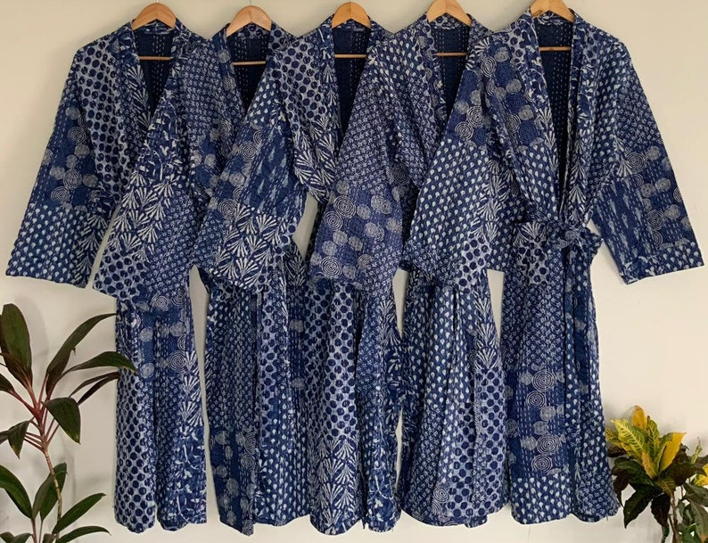 May include: Five indigo blue robes with white patterned designs hang on wooden hangers. The robes feature various patterns, including polka dots, floral motifs, and geometric shapes. Each robe has a matching belt.
