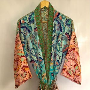 May include: A vibrant, patterned robe with a paisley design. The robe features a mix of blue, green, pink, and orange hues. It is hanging on a wooden hanger, showcasing its flowing fabric and intricate details. The robe is tied at the waist with a matching belt.