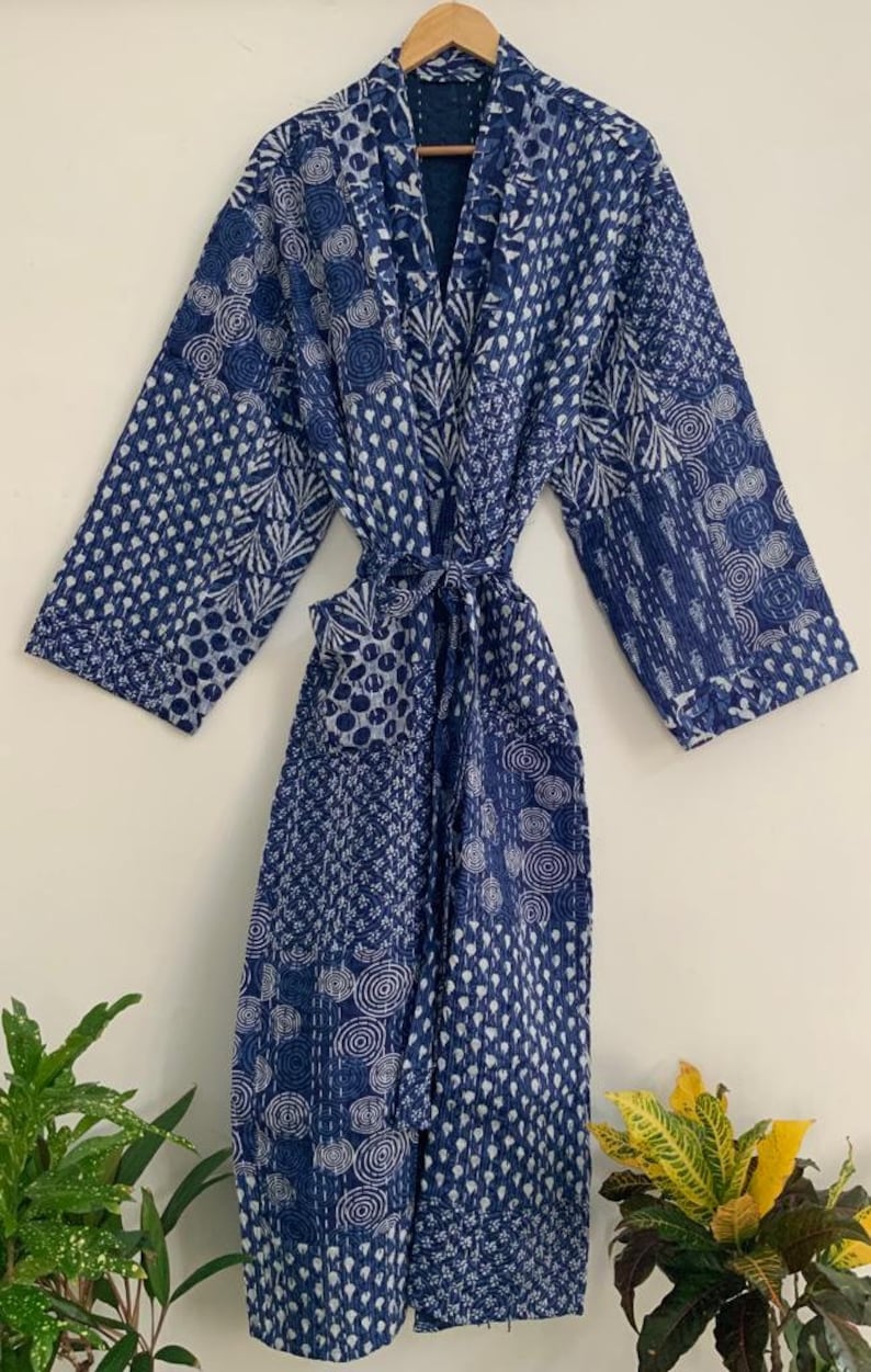 May include: A blue and white patterned robe with a tie waist. The robe is made of a patchwork design with various geometric and floral patterns. The robe is hanging on a wooden hanger.