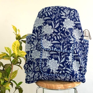 May include: A navy blue backpack with a white floral design. The backpack features a front pocket and a side pocket, and is placed on a wooden stool. A stainless steel water bottle is in the side pocket. The backpack is made of quilted fabric.