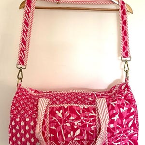 May include: A pink and white patterned duffel bag with a long shoulder strap. The bag has a quilted design and is made of cotton.