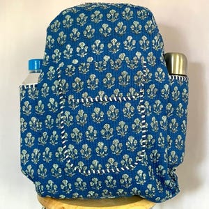 Op de afbeelding: Blauwe rugzak met een bloemenpatroon. De rugzak heeft twee zijvakken, één met een waterfles en de andere met een roestvrijstalen thermoskan. De rugzak heeft witte en marineblauwe afwerking.