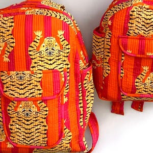 May include: Two quilted backpacks with a vibrant tiger print. The backpacks feature a repeating pattern of orange and pink vertical stripes with yellow tigers. Each backpack has a front pocket and adjustable straps.