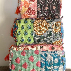 May include: Three quilted cosmetic bags in a stack. Each bag has a patchwork design with floral and geometric patterns in orange, pink, green, blue, and purple. The bags have orange tassels. The bags are made of a quilted fabric.