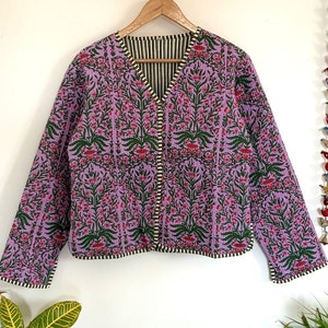 Puede incluir: Una chaqueta acolchada de color morado con estampado floral y forro a rayas verde y blanco. La chaqueta tiene cuello en V y cierre de botón.