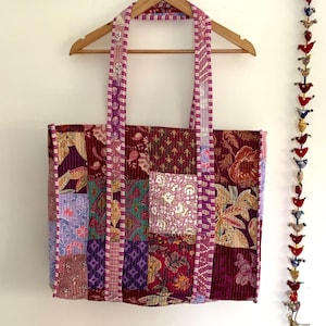 Peut inclure: Un sac cabas en patchwork avec une palette de couleurs rose et violette. Le sac est fait de tissu matelassé avec une variété de motifs floraux et géométriques. Le sac a deux longues anses et est suspendu à un cintre en bois.