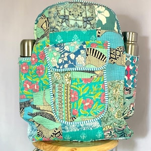May include: A patchwork backpack with a variety of floral and geometric patterns in shades of teal, blue, and pink. The backpack has two side pockets for water bottles and a front pocket. The bag is sitting on a wooden stool.