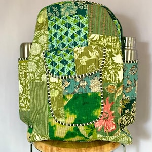 Può includere: Zaino patchwork verde con vari motivi floreali e geometrici. Presenta due tasche laterali per bottiglie d'acqua e una tasca frontale con un dettaglio floreale rosa. Lo zaino è appoggiato su uno sgabello di legno.