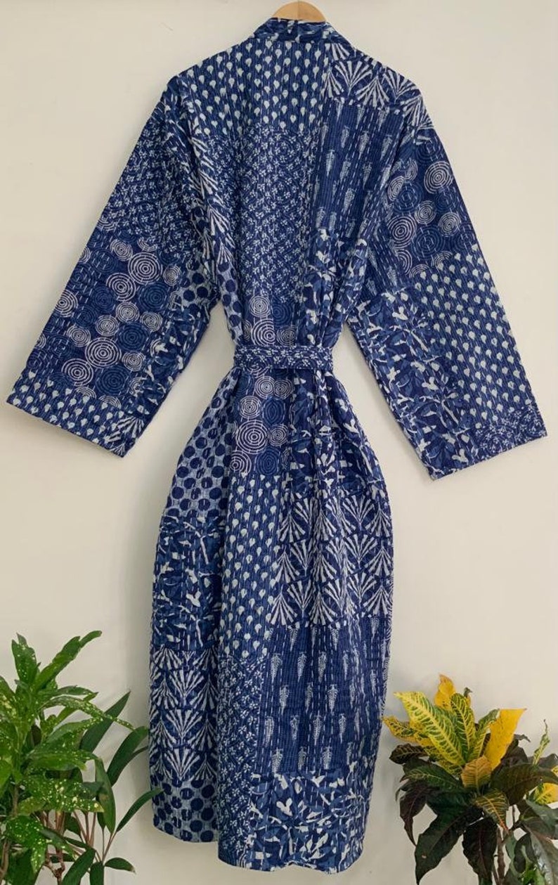May include: A long, indigo blue robe with a patchwork design. The robe features a variety of patterns, including circles, dots, and leaf-like shapes, in white against the blue background. It has long sleeves and a matching belt.