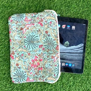 Peut inclure: Une housse de tablette à motifs floraux dans les tons de turquoise, rose et marron, avec une fermeture éclair blanche. La housse est posée à côté d'une tablette noire sur de l'herbe verte. Le motif floral présente des fleurs et des feuilles stylisées.