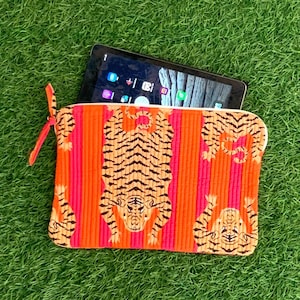 Peut inclure: Pochette rectangulaire orange et rose à fermeture éclair avec un imprimé tigre. Une tablette noire est visible à l'intérieur. Le motif tigre est noir et beige. La pochette a une petite sangle orange attachée à la fermeture éclair.
