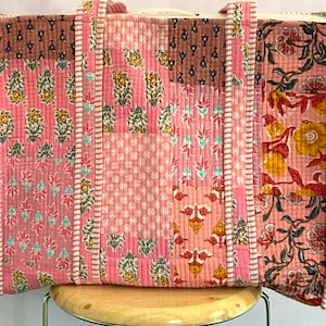 May include: A large, quilted tote bag with a patchwork design. The bag features various floral patterns in shades of pink, brown, and yellow. The handles and edges are trimmed with a white and pink striped fabric. The bag is sitting on a wooden stool.
