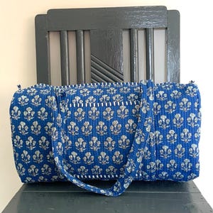 May include: A blue quilted travel bag with a white and light blue floral design. The bag has a rectangular shape, a zip closure, and a long strap. The bag is on a dark grey chair.