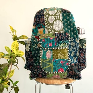 May include: A patchwork quilted backpack with a green, blue and black floral pattern. The backpack has a large pocket on the front and a smaller pocket on the side. A water bottle is in the side pocket.