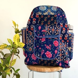 Puede incluir: Una mochila azul marino con un estampado floral y geométrico. La mochila tiene un bolsillo frontal con flores rosas y ribete blanco. Tiene dos bolsillos laterales y está sobre un taburete de madera. Dos botellas de agua de acero inoxidable están en un bolsillo lateral.