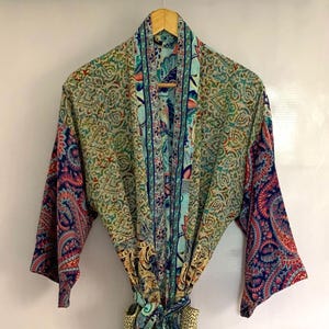 May include: A colorful, patterned robe with a paisley design. The robe features a mix of colors, including blue, green, red, and yellow. It is hanging on a wooden hanger against a white background. The robe has long sleeves and a tie closure.