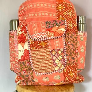 May include: A coral patchwork backpack with floral and geometric patterns. It has two side pockets, each holding a stainless steel water bottle. The bag is on a wooden stool. The backpack's design incorporates peach and cream hues.
