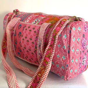 May include: Pink quilted duffel bag with floral print and white stripes. The bag has two straps and a top handle.