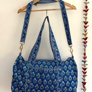 May include: A blue tote bag with a floral pattern and white stitching. The bag has a matching adjustable shoulder strap and gold-colored metal hardware. The bag is hanging from a wooden hanger.