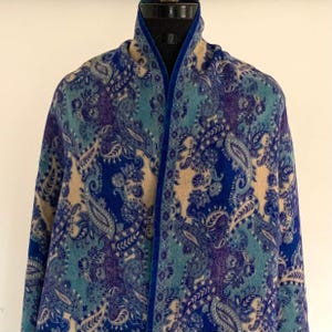 Könnte beinhalten: Ein Paisley-Schal in Blau-, Türkis- und Lilatönen. Der Schal wird auf einer Schaufensterpuppe präsentiert, die das detaillierte Design hervorhebt. Die Kanten sind mit einem dunkelblauen, samtartigen Material verziert. Das Muster weist florale und geschwungene Motive auf.