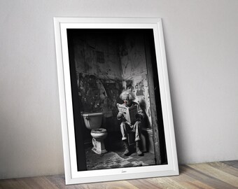 May include: Black and white framed print of a person reading a newspaper while sitting on a toilet. The image is in a bathroom with peeling paint and a vintage toilet. The person has white hair and a red nose.