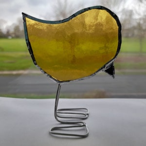 May include: A gold stained glass bird with a black outline, standing on a silver wire stand.