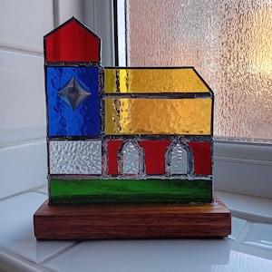 May include: A stained glass window design of a building with a red roof, blue, yellow, white, and green glass panels. The window is mounted on a wooden base.