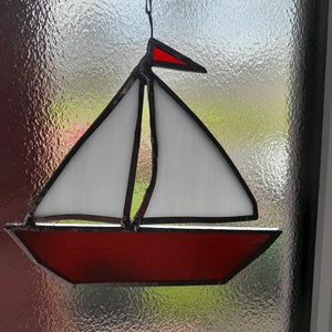 Puede incluir: Un velero de vitral con una vela y un casco rojos. El velero está colgado de una ventana.