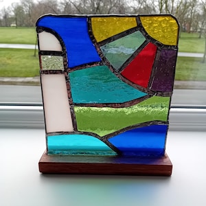 May include: A colourful stained glass panel with a geometric design. The panel features blue, green, yellow, red, and purple glass pieces. It is mounted on a rectangular wooden base. The glass is textured and the panel is placed in front of a window.