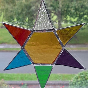 May include: A stained glass star ornament with six points. The star is made of six different coloured glass pieces: red, orange, yellow, green, blue, and purple. The star is hanging from a white string.