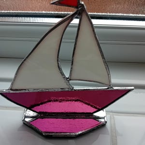 May include: Stained glass sailboat with white sails, a magenta hull, and a red pennant. The decorative nautical piece is made with lead solder and sits on a matching magenta base. A unique home decor item.