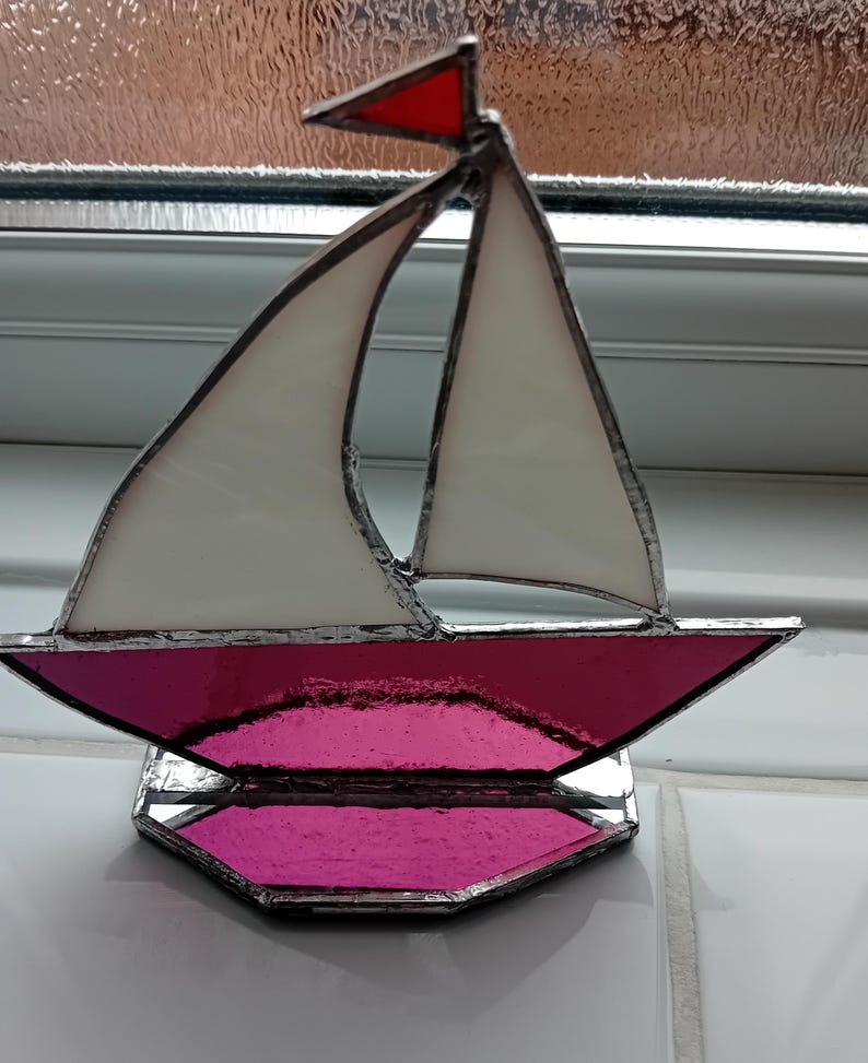 May include: Stained glass sailboat with white sails, a magenta hull, and a red pennant. The decorative nautical piece is made with lead solder and sits on a matching magenta base. A unique home decor item.