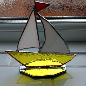 May include: A stained glass sailboat ornament. It features a yellow hull, white sails, and a red pennant. The boat sits on a clear, reflective base. The glass has a textured finish. The background is a neutral colour.