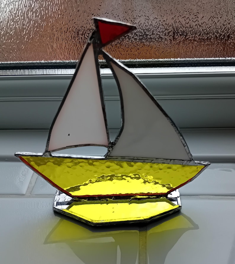May include: A stained glass sailboat ornament. It features a yellow hull, white sails, and a red pennant. The boat sits on a clear, reflective base. The glass has a textured finish. The background is a neutral colour.