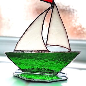 May include: A stained glass sailboat with a red flag, white sails, and a textured green hull. The boat sits on a clear, faceted base. The sailboat is a decorative nautical item.